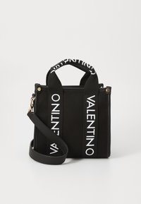 Bolso tote negro de material de lona, con dos asas superiores y correa de hombro removible. Presenta una correa con el texto "VALENTINO" y acentos en dorado.