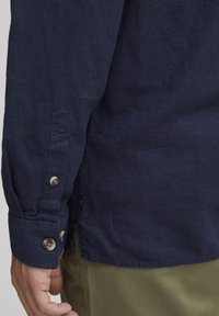 Camisa de manga larga en color marino, hecha de lino, con puños abotonados y un dobladillo recto. La tela tiene una textura suave con sutiles detalles de costura.