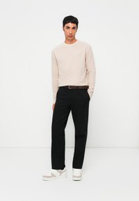 Man standing with hands in pockets wearing beige sweater, black pants, brown belt, and white sneakers against plain white background.