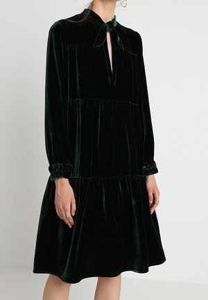 Langærmet, mørkegrøn fløjls kjole med lagdelt nederdel, elastiske manchetter og en lille sløjfe ved halsudskæringen.