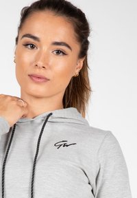 Grijze hooded sweatshirt met zwarte koorden en een zwart logo op de borst. De stof lijkt zacht met een gladde textuur.