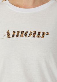 T-shirt en coton blanc avec le mot "Amour" dans un design imprimé léopard. Le t-shirt a un col rond et des manches courtes.
