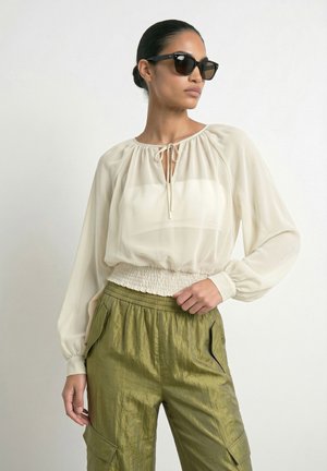 Mujer con el cabello oscuro recogido en un moño, lleva gafas de sol negras, blusa crema de manga larga con lazo al cuello y pantalones verde oliva brillantes con bolsillos.