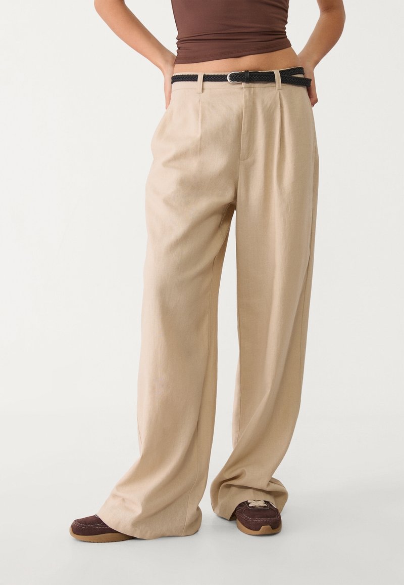 Personne portant un pantalon beige à jambes larges avec une ceinture noire tressée, un haut marron et des chaussures décontractées marron sur un fond clair uni.