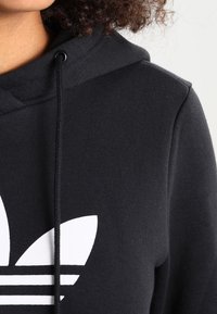 Svart hoodie med en vit trefoil-logotyp, med dragsko i huvan, mjuk textur och avslappnad passform.