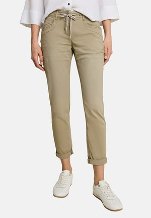 Frau trägt hochgekrempelte beige Hose mit Fronttaschen und weißen Sneakers, kombiniert mit einem weißen Hemd mit Knöpfen und weiten Ärmeln.