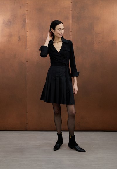 Čierne zavinovacie šaty s hlbokým výstrihom do V, dlhými rukávmi a plisovanou sukňou. Dopĺňajú ich čierne členkové čižmy so zlatými akcentmi. Hladká textúra.