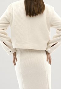 Valkoinen tekstuurinen lyhyt takki, jossa on pitkät hihat ja napilla kiinnitettävät hihansuut, yhdistettynä korkeavyötäröiseen hameeseen. Yksinkertaiset, puhtaat linjat.