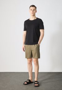 Camiseta negra de manga corta, pantalones cortos caqui y sandalias negras con hebillas. La camiseta tiene un cuello redondo y un corte relajado; los pantalones cortos son de longitud hasta la rodilla.