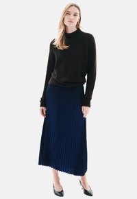 Frau trägt einen dunkelbraunen Langarmpullover, einen blauen plissierten Midirock und schwarze spitze High Heels, steht vor einem einfarbigen Hintergrund.