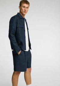 Marineblå jakke med knapper og to brystlommer, parret med matchende shorts. Hvid skjorte under, tekstureret stof, afslappet pasform.