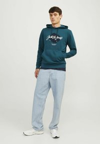 Sudadera color azul verdoso con texto gráfico en blanco, bolsillo canguro y capucha con cordón, combinada con jeans de corte ancho de color azul claro y zapatillas blancas.