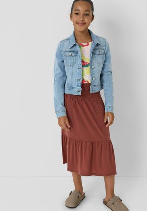 Junges Mädchen trägt eine hellblaue Jeansjacke, ein bunt gemustertes Oberteil, einen rostfarbenen Rock und beige Slipper, steht vor einer schlichten Wand.