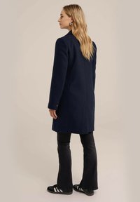 Tamsiai mėlynas vilnonis paltas, pritaikytas, ilgomis rankovėmis ir švaria siluetu, derinamas su juodomis plačiomis džinsomis ir juodais sportbačiais.