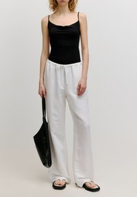 Schwarzer, figurbetonter Bodysuit, weiße Leinen-Hosen mit weitem Bein und elastischem Bund, schwarze flache Sandalen und eine schwarze Tote-Tasche.