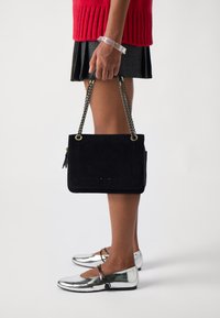 Svart mockaväska med en strukturerad design, guldaccessoarer och kedjesnor. Bärs med en röd stickad topp och en grå plisserad kjol, matchad med silverfärgade skor.