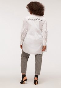 Camicia bianca a maniche lunghe con testo ricamato "non guardare indietro" sulla schiena. Abbinata a pantaloni grigi e sandali neri con tacco.