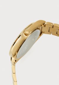 Montre en métal doré avec un cadran texturé, boîtier rond et bracelet à maillons ajustable. Elle est dotée d'une couronne et d'un fond lisse argenté.