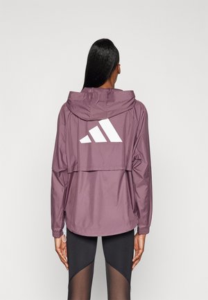 Woman wearing a purple hooded jacket with a large white Adidas logo on the back, paired with black leggings with sheer panels.