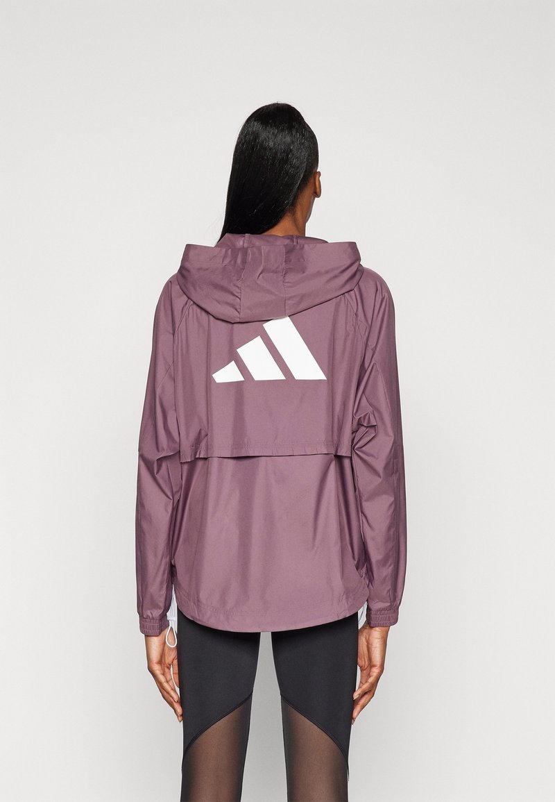 Femme portant une veste à capuche violette avec un grand logo Adidas blanc dans le dos, associée à des leggings noirs avec des panneaux transparents.