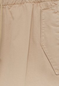 Gros plan sur un pantalon en tissu beige avec une ceinture élastique et une poche latérale, présentant un détail de couture visible.