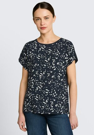 Frau mit dunklen Haaren in einem niedrigen Dutt, die eine kurzärmelige marineblaue Bluse mit weißen abstrakten Flecken und blaue Jeans trägt, steht vor einem schlichten Hintergrund.