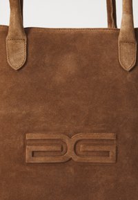 Bruine suede tote bag met reliëflogo. Soepele textuur met stevige handvatten. Eenvoudig ontwerp, met de focus op materiaal en vakmanschap.