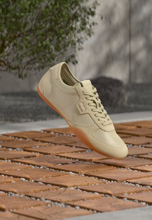 Beiger Schnürsneaker mit orangefarbener Sohle, der über hölzernen Fliesen im Freien schwebt, im Hintergrund eine Blattpflanze und eine Steinmauer.