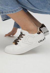 Bijele kožne tenisice s uzorkom leoparda na vezicama i teksturiranim petnim dijelom. Sadrže "LOLA CASA" branding sa strane i perforirane dizajnerske elemente.