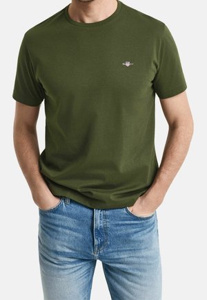 Homme portant un t-shirt uni vert olive à manches courtes avec un petit logo sur la poitrine et un jean bleu clair, mains dans les poches.