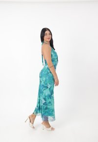 Abito senza maniche lungo fino al pavimento in teal con motivi floreali. Presenta una schiena annodata e una silhouette fluida, abbinato a sandali bianchi con tacco.