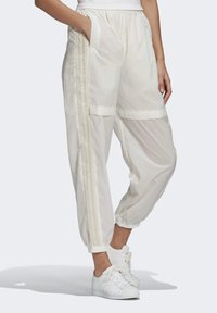 Vita lätta träningbyxor med resårmanchetter och tre vertikala ränder på sidan, bärs med vita sneakers och handen i fickan.