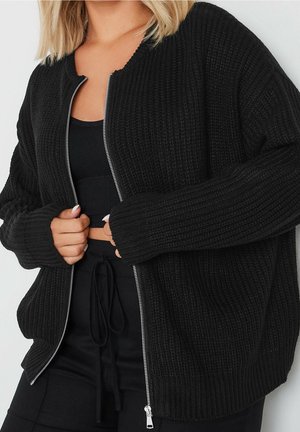 Femme portant un cardigan noir à fermeture éclair en maille côtelée, par-dessus un crop top noir et un pantalon noir à cordon de serrage.