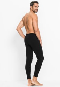 Schwarze Leggings aus dehnbarem Stoff, enganliegendes Design, verjüngen sich an den Knöcheln. Das Modell zeigt eine Rückansicht vor einem weißen Hintergrund.