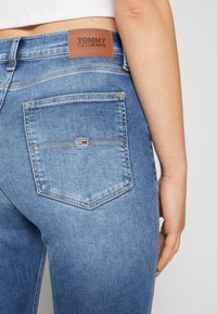 Blå denimjeans med ljus tvätt, med en brun läderlapp märkt "TOMMY JEANS" och en ficka med en liten röd, vit och blå logotyp.