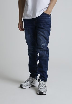 Person iført mørkeblå jeans med let slid og hvide sneakers, stående mod en ensartet lys grå baggrund.