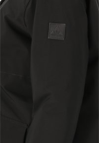 Svart jacktyg med en slät yta. Har en rektangulär svart logotypflicta som visar en bergdesign på överarmen.