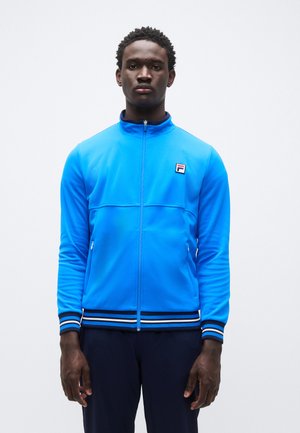 Kék sportos cipzáras kabát sima anyagból, fekete és fehér csíkozott díszítésekkel az ujjvégeken és az alsó szegélyen, a mellkason egy kis logóval.