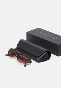 Gafas de sol con tintado marrón y forma redondeada, estuche de transporte negro texturizado y una caja negra con logotipo en relieve.