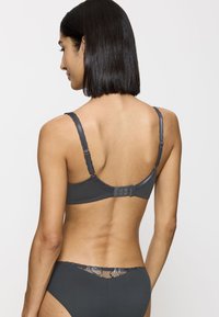 Nainen, jolla on suorat mustat hiukset, pukeutunut tummanharmaaseen ribbikaulushousut ja vastaavat alushousut, joissa on pitsiyksityiskohtia. Hänet näkyy selkäpuolelta valkoista taustaa vasten.