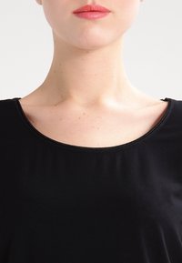 Černý top se širokým, zaobleným výstřihem a krátkými rukávy. Látka působí hladce, s jemným leskem a minimem švů.