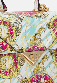 Lähikuva värikkäästä käsilaukkukankaasta, jossa on koristeellisia kultakuvioita, vaaleanpunaisia ja turkooseja kuvioita sekä kultainen kolmionmuotoinen solki.