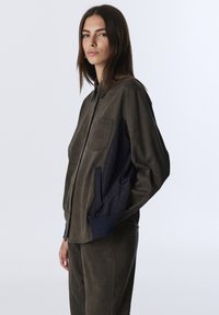 Kobieta ubrana w brązową koszulę zapinaną na guziki z czarnymi panelami bocznymi oraz pasującymi spodniami, stojąca na tle jasnego, jednolitego tła.