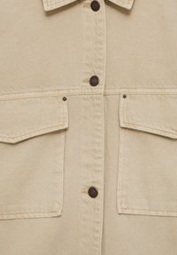 Veste en denim beige avec deux poches avant à rabat et des boutons-pression en métal foncé au centre et sur les rabats des poches.