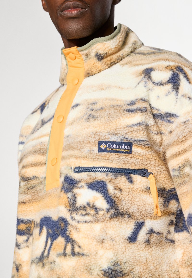 Pullover in pile di colore crema con motivi astratti blu e gialli. Presenta una chiusura a metà cerniera, bottoni arancioni e una tasca con cerniera sul petto.