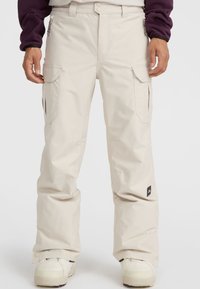 Beige snow pants featuring a loose fit, multiple side pockets, and reinforced stitching. Material appears water-resistant with a smooth texture.