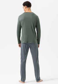 Man die blootsvoets staat met zijn rug naar ons gekeerd, met een lange mouw olijfgroene top en marineblauwe gestreepte pyjamabroek tegen een witte achtergrond.