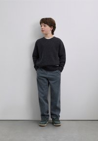 Pull en maille texturée foncée, pantalon en velours côtelé gris et baskets vertes. L'enfant se tient les mains dans les poches devant un fond neutre.