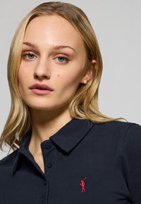 Polo shirt marine avec un col à boutons, ornée d'un logo brodé rouge d'un golfeur sur la poitrine gauche. Texture lisse et design ajusté.