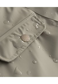 Wasserabweisender beiger Stoff mit sichtbaren Wassertropfen. Verfügt über eine schräg angebrachte Fronttasche und ein kreisförmiges Metall-Logo.